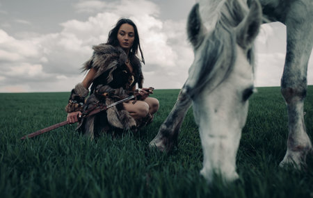 Woman sits on meadow in image of ancient warrior amazon armed by sword near grazing horse.の写真素材