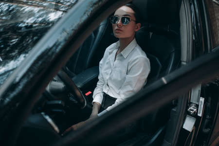 Beautiful young woman sits behind the wheel in black modern car. View through opened door.の写真素材