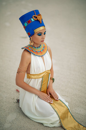 Young woman in image of ancient egyptian queen Nefertiti sits in sandy desert.の写真素材
