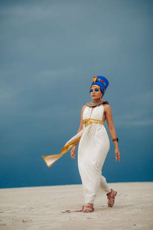 Young woman in image of ancient egyptian queen Nefertiti walks through sandy desert against background of blue sky.の写真素材