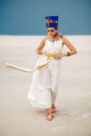 Young woman in image of ancient egyptian queen Nefertiti walks through sandy desert against...