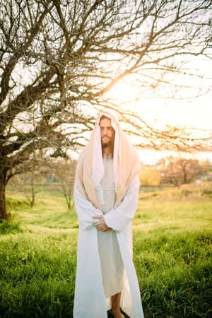 Jesus Christ standing in meadow clothed in his traditional white robe against tree and sunset background.の写真素材
