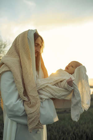 Saint Joseph holds baby Jesus in his arms and looks to him with love against meadow and sky...