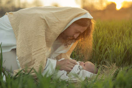 Saint Joseph bents over baby Jesus lying on grass and looks to him with love at sunset.の写真素材