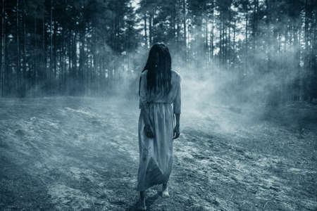 Girl with long black hair in image of scary ghost zombie walks among dark forest against background of smoke.の写真素材