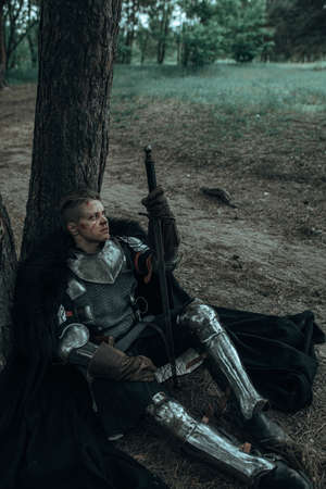 Medieval tired warrior knight in chain mail armor and black cape sits and rests among forest under...