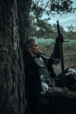 Medieval tired warrior knight in chain mail armor sits and rests among forest under tree with sword in his hand.の写真素材