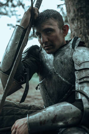Medieval tired warrior knight in chain mail armor sits and rests among forest under tree with sword in his hand.の写真素材