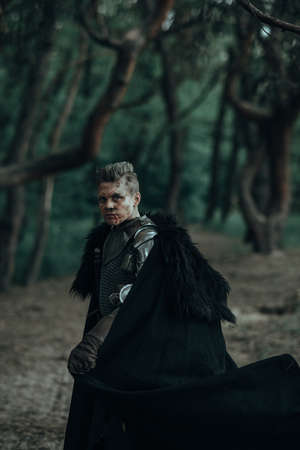 Medieval knight in chain mail armor and black cape stands among forest.の写真素材