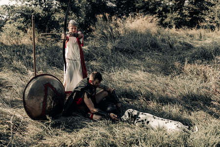 Ancient Spartan warrior in battle dress and woman archer rest with dog among meadow next to shield...