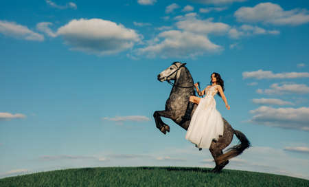 Bride in long white dress sits horseback on rearing horse against background of sky and clouds. With copy space.の写真素材