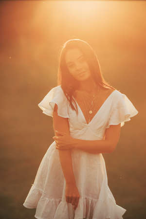 Young joyful woman in white dress stands in meadow at sunset on summer day. Backlight. Closeup.の写真素材