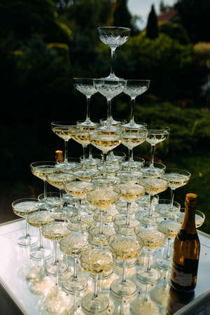 Pyramid of wine glasses with champagne is on table covered with white tablecloth at wedding banquet against the backdrop of lawn and garden.の写真素材