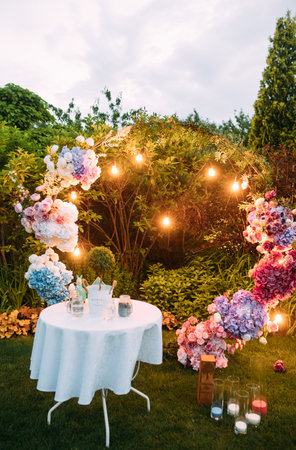 Stylish wedding arch decorated fresh flowers with glowing garland and table with white tablecloth are in the beautiful landscaping garden among lawn.の写真素材