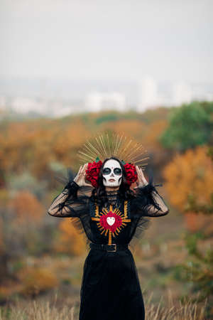 Young woman with sugar skull makeup and red roses dressed in black costume of death as Santa Muerte is against background of autumn forest. Day of the Dead or Halloween concept.の写真素材