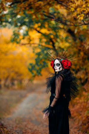 Young woman with sugar skull makeup and red roses dressed in black costume of death as Santa Muerte...