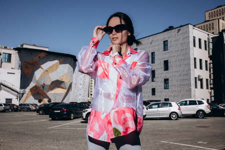 Stylish young woman in sunglasses stands dressed in colorful transparent blouse and white shorts against cars and building background.の写真素材