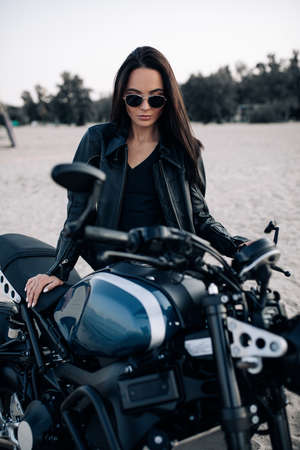 Young woman stands near black motorbike in sunglasses and leather jacket against sandy beach background.の写真素材
