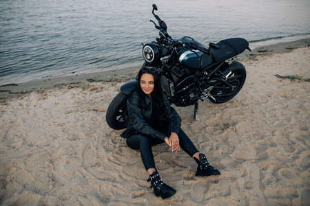 Young woman sits on sand near black motorbike in leather jacket and pants on beach near river coast.の写真素材