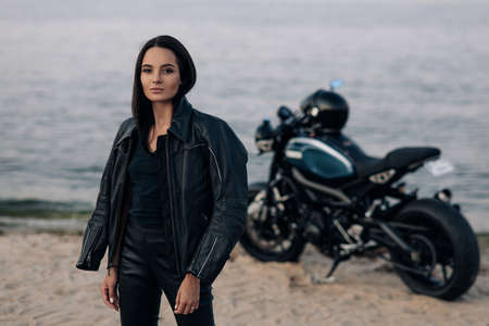 Young woman stands next to black motorbike in leather jacket and pants on sandy beach near coast of...