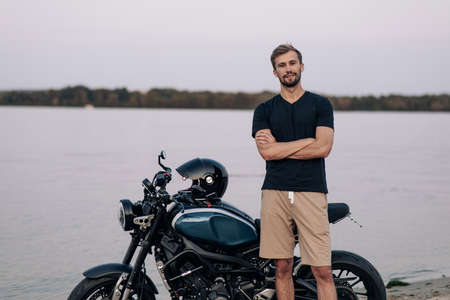 Young man stands next to black motorbike on sandy beach near coast of river.の写真素材