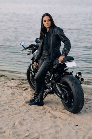 Young woman stands next to black motorbike in leather jacket and pants on sandy beach near coast of...