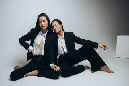 Two young brunette women pose in studio sitting dressed in black suits.の写真素材