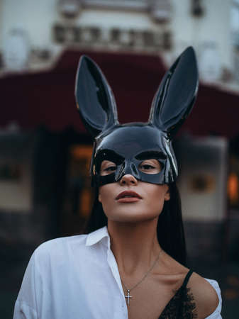 Portrait of young beautiful sexy woman in carnival black mask of Easter bunny and white shirt against city background.の写真素材