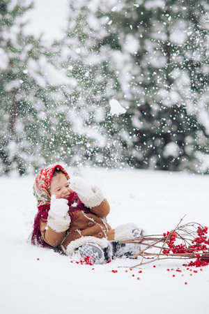 Beautiful little child girl plays in snowy forest during snowfall. She dressed in the old Russian style in red headscarf.の写真素材