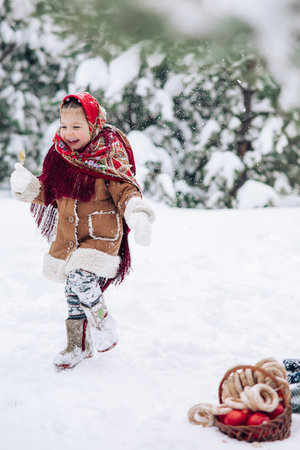 Beautiful little child girl plays in snowy forest near basket with apples and bagels. She dressed in the old Russian style in red headscarf.の写真素材