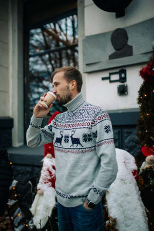 Young man stands near building and drinks hot coffee in street decorated for Christmas.の写真素材