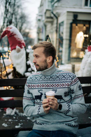 Young man sits on bench and drinks hot coffee in snowy street decorated for Christmas. closeup.の写真素材