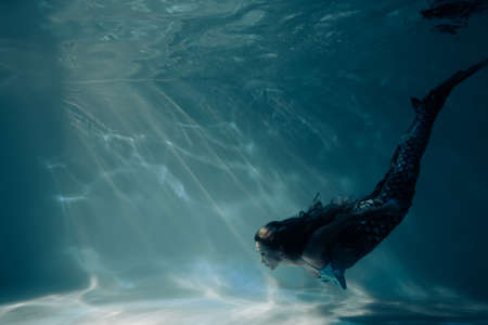 Beautiful young woman with mermaid tail swims and dives under sunlit deep water.の写真素材