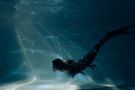 Beautiful young woman with mermaid tail swims and dives under sunlit deep water.