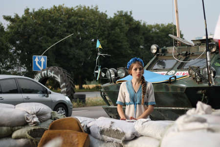 Young ukrainian woman in traditional national dress stands on roadblock against background of armored personnel carrier and sandbags. Concept of Russian military invasion in Ukraine. War in Ukraine and Europe.の写真素材