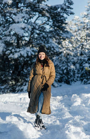 Happy young smiling woman walks in forest among snow-covered pine trees in sunny winter day.