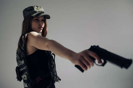 Young woman in uniform stands and aims with handgun in her hand against gray background.の写真素材