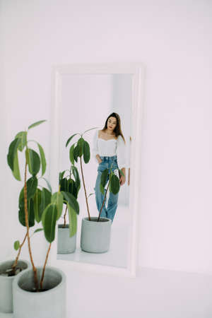 Reflection in mirror of young brunette woman standing next to flowerpot with ficus houseplant and looking on herself.の写真素材