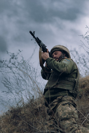 Ukrainian soldier aims from  assault rifle to russian enemy against background of hill with dry grass and sky. Concept of Russian military invasion in Ukraine. War in Ukraine and Europe.の写真素材