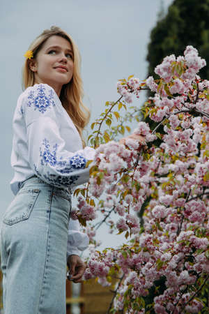 Young blonde woman walks in garden dressed in Ukrainian embroidered shirt and enjoys by blooming sakura trees.の写真素材
