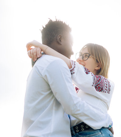 Interracial couple embraces against sky background dressed in Ukrainian traditional ethnic...