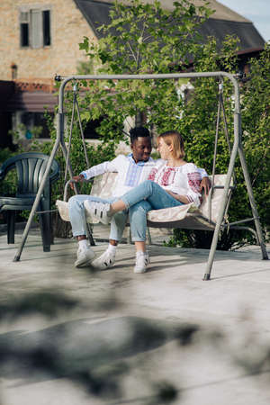 Interracial couple sits on bench swing in garden dressed in Ukrainian traditional ethnic embroidered shirts. Concept of love relationships and unity between different human races.の写真素材