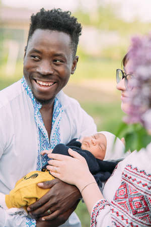Happy interracial family dressed in Ukrainian national embroidered shirts holds their newborn baby....