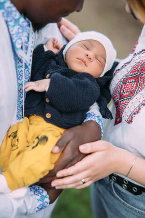 Happy interracial family dressed in Ukrainian national embroidered shirts holds their newborn baby. Concept of interracial family and unity between different human races. closeup.の写真素材