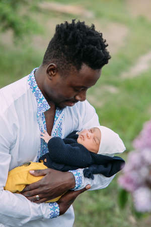 Happy african man dressed in Ukrainian national embroidered shirt holds his mulatto newborn baby...