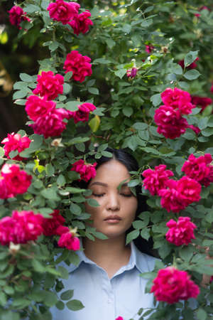 Portrait of young asian woman with closed eyes enjoying by blooming red climbing rose in garden.の写真素材