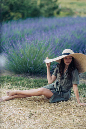Young woman in wide-brimmed hat sits on straw and rests among lavender field enjoying by blooming.の写真素材