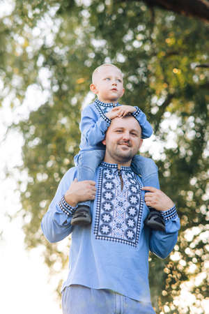 Happy father carries his son on his shoulders during walk on park. They are dressed in Ukrainian national embroidered shirts.の写真素材