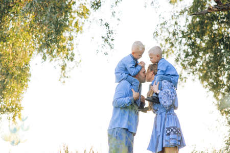 Happy parents carry their two twins sons on their shoulders and kiss each other during walk on meadow. They are dressed in Ukrainian national embroidered shirts.の写真素材