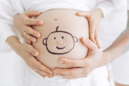 Couple parents touch pregnant belly by their hands with drawn smiling baby face on it . Concept of love and care during pregnancy, expecting and born baby.の写真素材